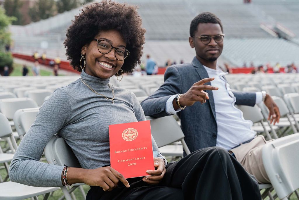 Two family members attending Boston University's 147th Commencement sit on Nickerson Field and pose for a photo holding the Red Book program for the ceremony.