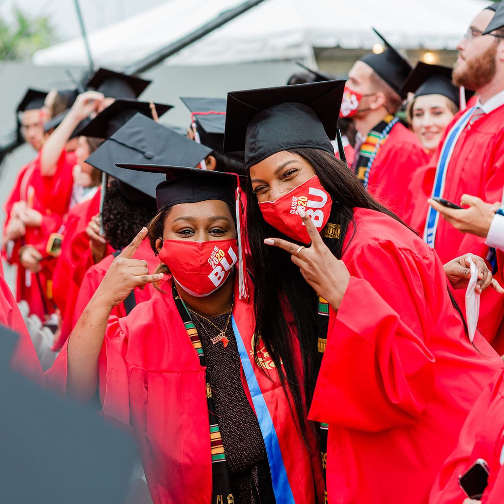 Photo of two young Black women at 2020 Commencement ceremony. They were their black caps and red gowns and make the letter V with their hands. They smile from underneath their red masks.