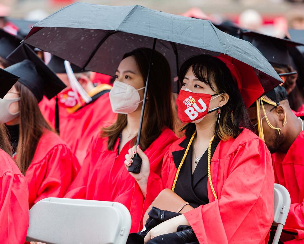 Two BU Class of 2020 graduates wearing face coverings hold a black umbrella over their head while watching the Commencement ceremony.