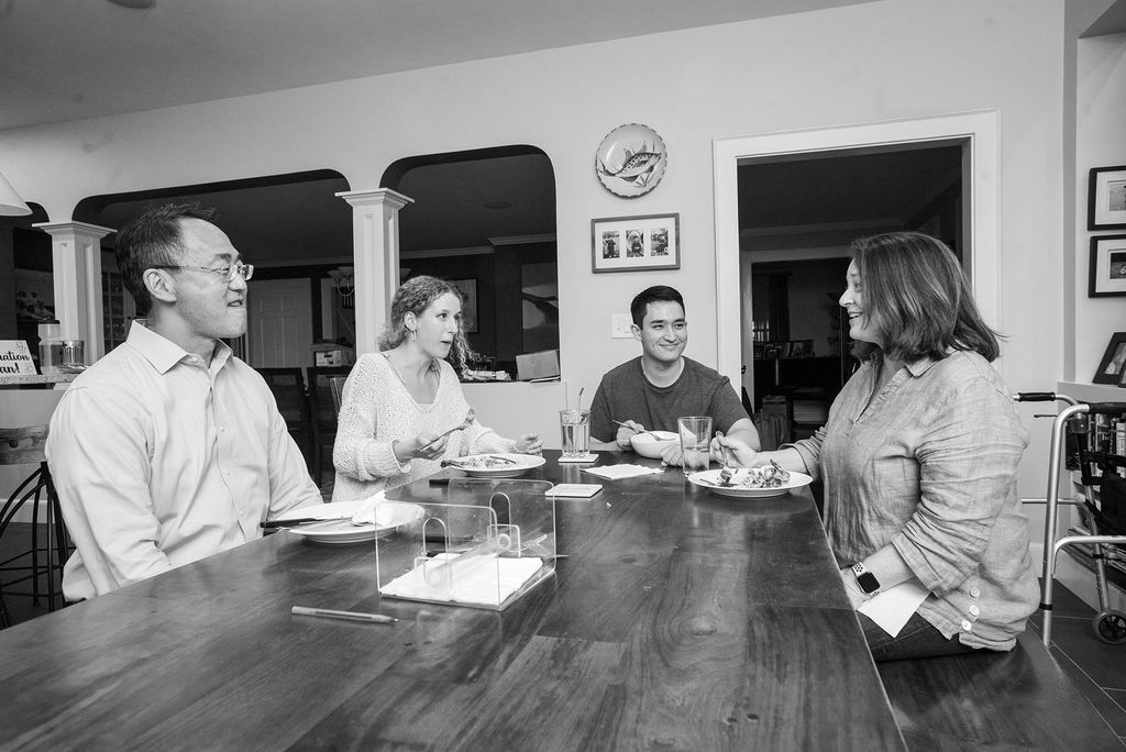 Black and white photo of Willis Wang, from left, Phoebe Bobola (CAS’20) , Ethan Wang (CAS’20) and Ethan’s mom, Teri Yahia at the end of the day in the Wang home September 28. They sit at a large wooden table and smile as they eat. A half wall opens up into the rest of the house behind them.