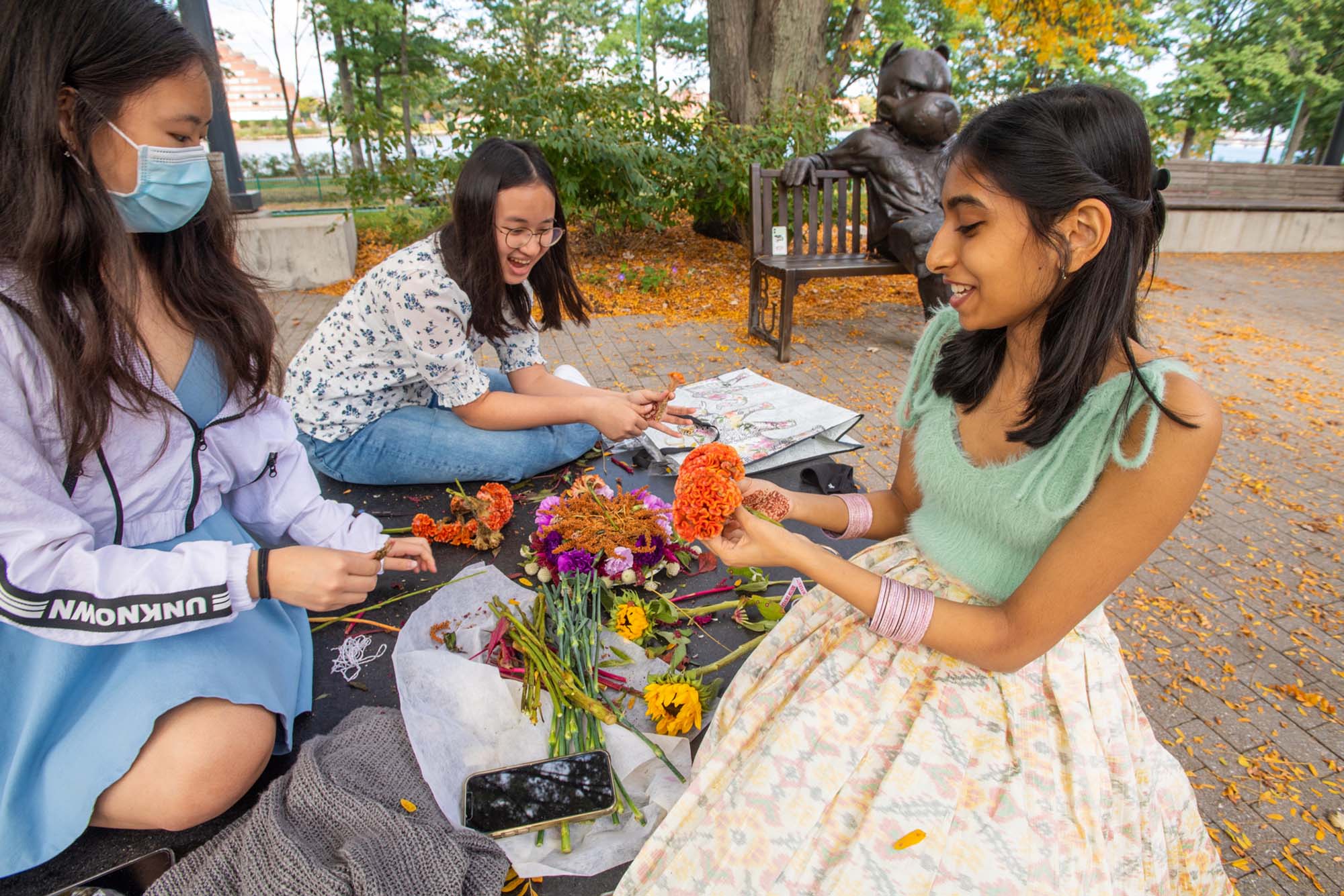 Photo of friends Heather Li (CFA’25), from left, Caitlyn Alysha Aryanto (Questrom’25), and Anvita Reddy (ENG’25) building a Bathukamma, a flower stack arranged with different seasonal flowers in seven concentric layers in the shape of temple gopuram. They sit on the ground near the Rhett bench amidst yellow, fallen leaves, and laugh joyfully.