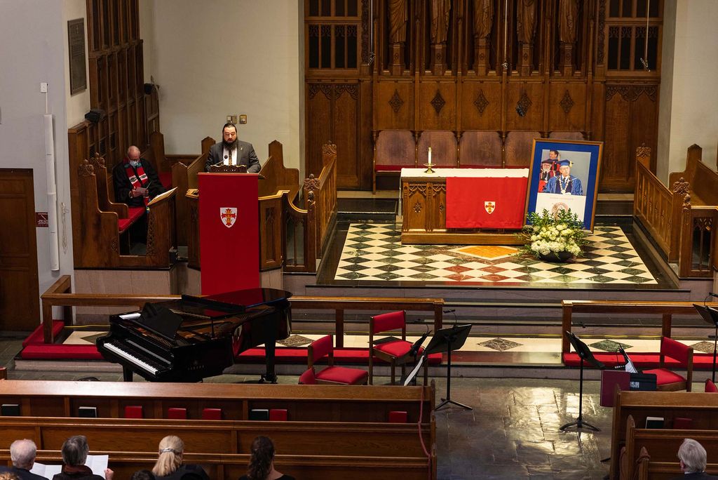 Photo taken during a Memorial for former BU president Jon Westling on Wednesday at March Chapel. At the podium at left, Westling's son Matthew Westling reads "Invective Against Swans," a poem by Wallace Stevens. Behind him, sits Reverend Hill. A photo of Westling leans against the altar at right. The heads of a few people sitting in the first pew are seen.