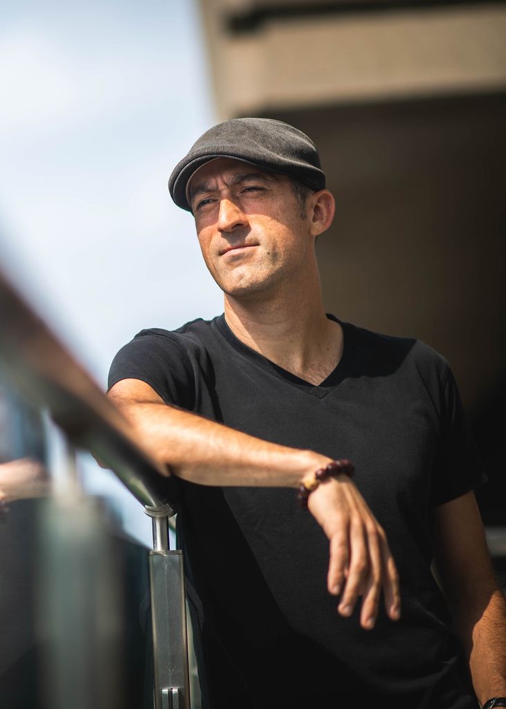 Photo of David Delmar Senties, wearing a gray hat, and a black t-shirt, as he leans across a clear balcony with a silver railing.