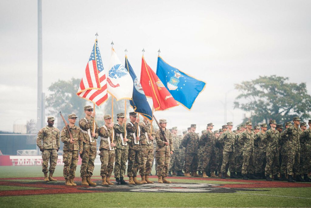 Photo of Pass and Review on Nickerson Field. From left to right, Commander Cadet Nathan Tadigiri, Rifleman- Cadet Kenneth Ziniti (Army), National Colors- Cadet Salem Adda-Berkane, Army Colors- Cadet Joseph Carey, Navy Colors- MIDN Sasha Wong Marine Corps, Colors-MIDN Ian Benitez-Rio Air Force Colors, Cadet Jacob Bresnahan Rifleman, Cadet Ju Young Kang (Army). They each wear camouflage uniforms and hold flags and rifles. Behind them, a larger group of ROTC members are seen.