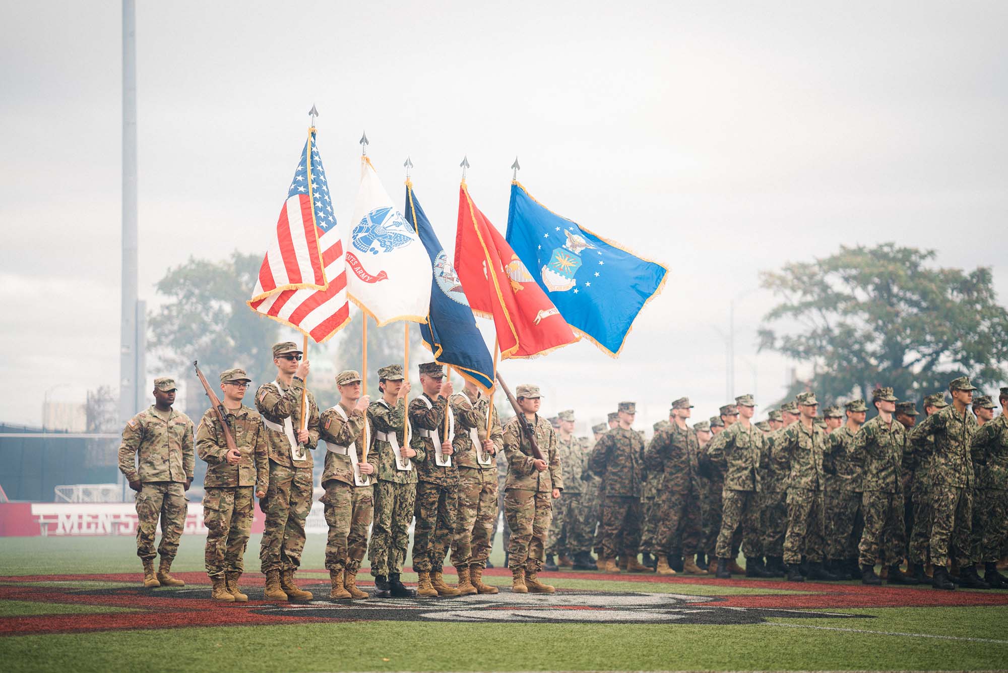 Photo of Pass-in-Review on Nickerson Field. From left to right, Commander Cadet Nathan Tadigiri, Rifleman- Cadet Kenneth Ziniti (Army), National Colors- Cadet Salem Adda-Berkane, Army Colors- Cadet Joseph Carey, Navy Colors- MIDN Sasha Wong Marine Corps, Colors-MIDN Ian Benitez-Rio Air Force Colors, Cadet Jacob Bresnahan Rifleman, Cadet Ju Young Kang (Army). They each wear camouflage uniforms and hold flags and rifles. Behind them, a larger group of ROTC members are seen.