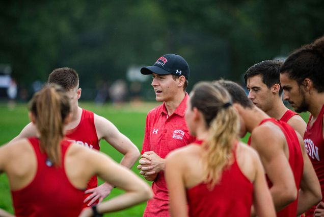 Photo of Jordan Carpenter, new associate head coach of cross country, with a blue Boston University hat on, speaking with cross country runners, dressed in red running uniforms.