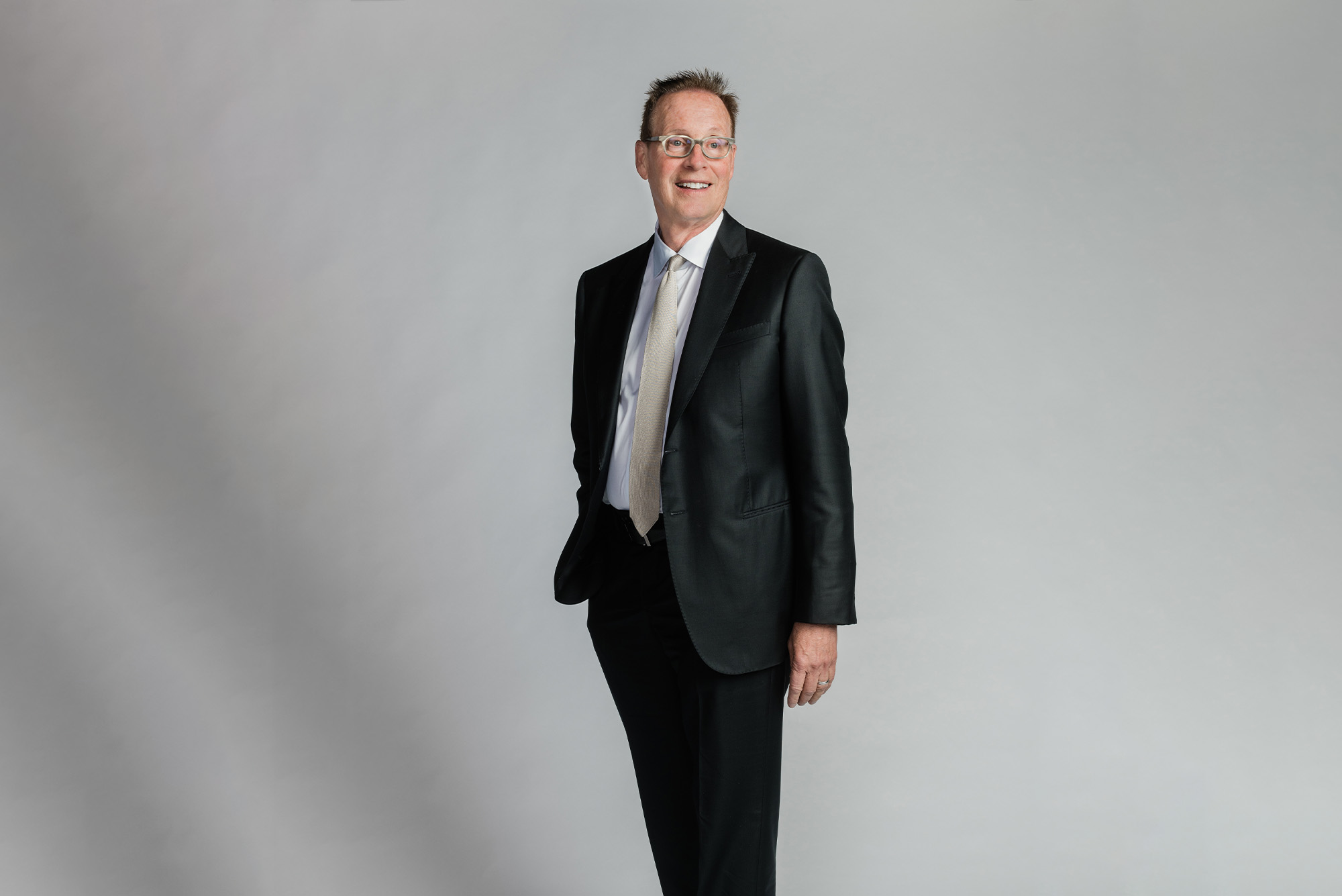 a studio photo of Boston University's Board of Trustees member Richard C. Shipley. He looks to the distance wearing a black suit in front of a grey background.
