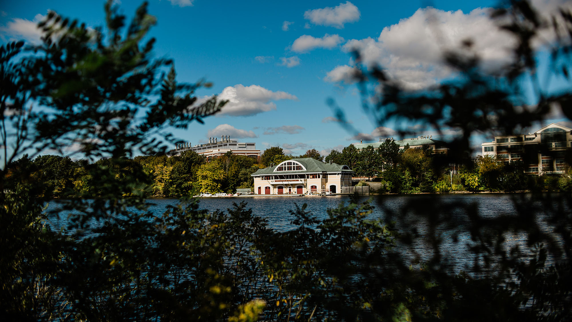 Photo of DeWolfe Boathouse taken from across the Charles River on a sunny fall day. Small clouds are seen in a brilliant, blue sky. The Boathouse is tan with a greenish gray roof.