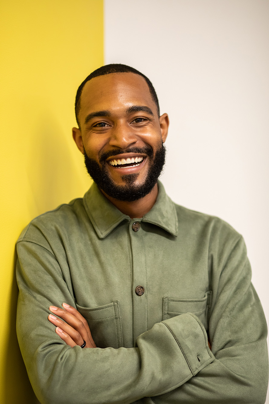 Photo of Jonathan Allen (LAW’19), a young Black man with a beard, leaning against a yellow wall. He wears a green jacket and smiles.