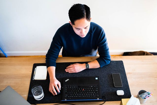 an overhead shot of Matt Bae working at his WFH desk. His desk features a dark grey felt mat, a Lenovo laptop that he works from, his cellphone and AirPods, a glass of water, a notepad, and a pen holder filled with multicolored Muji pens.