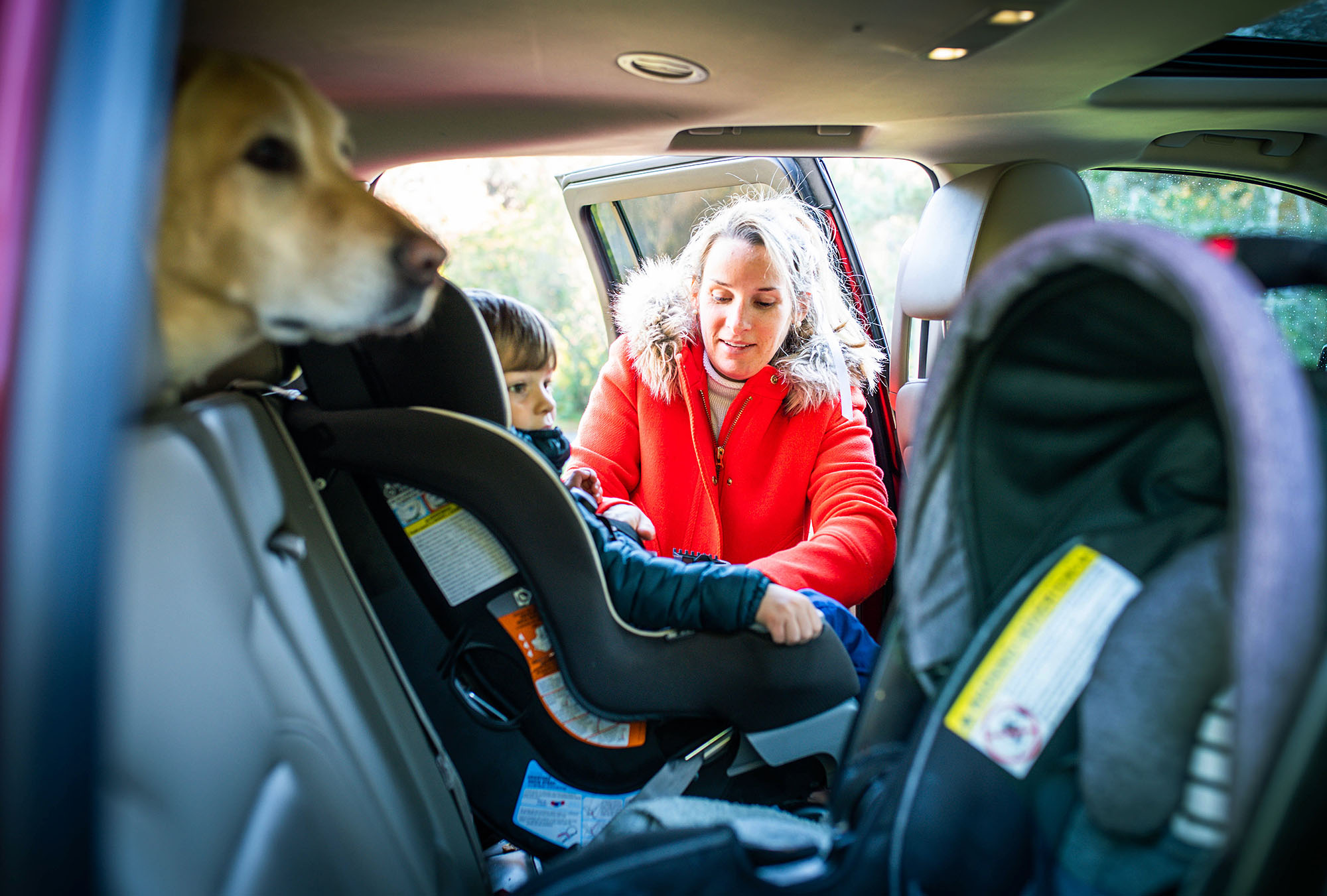 photo of Elizabeth Clancey as she wears a bright red coat and buckles her soon into his car seat. A labrador dog pokes its head from the back of the car into the shot.
