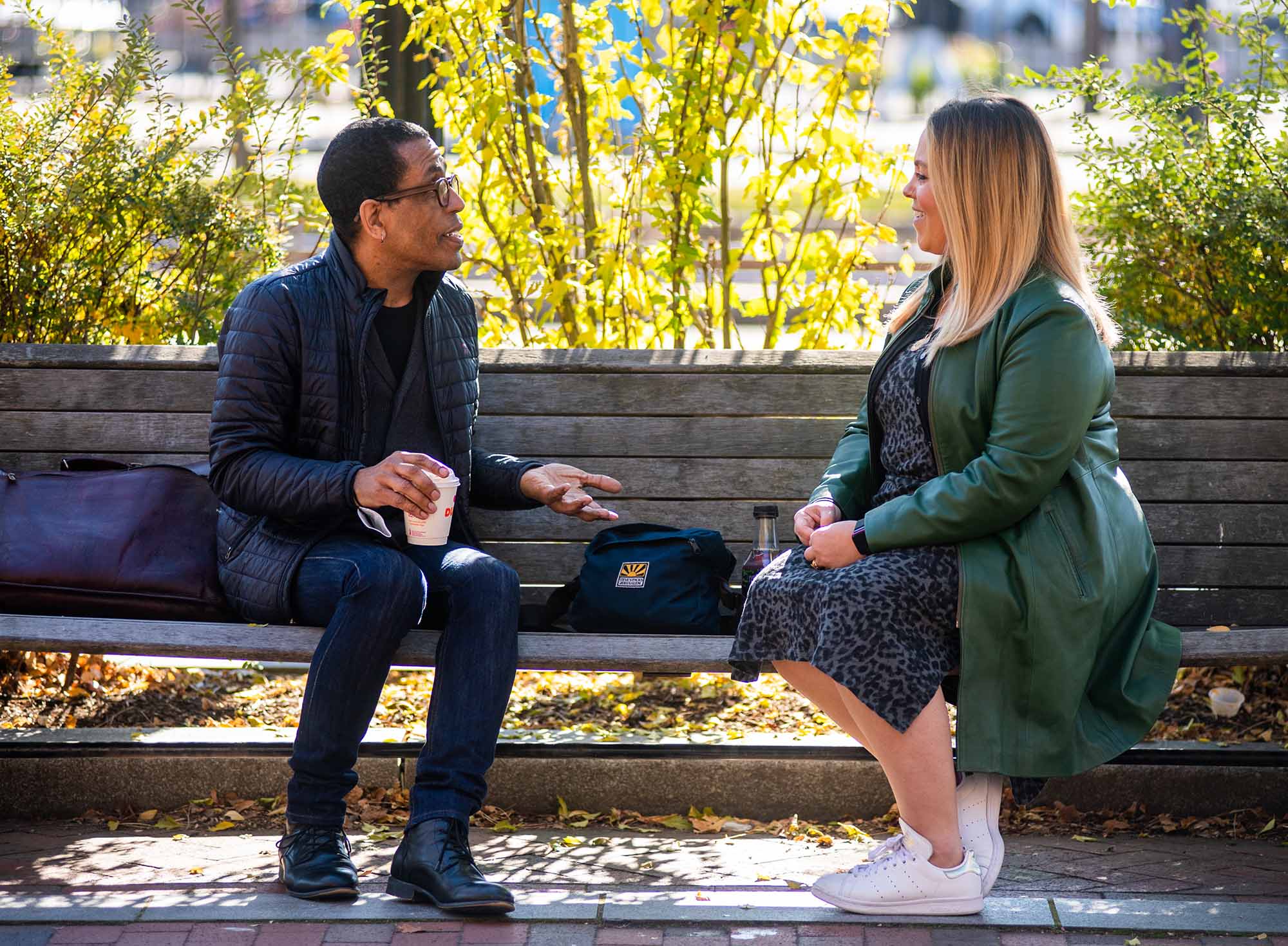 Candid photo of Ron Wheeler, left, chatting with Shira Megerman, senior legal librarian, on a wooden bench. Wheleer holds a cup of coffee as he chats. Background features yellow fall leaves.