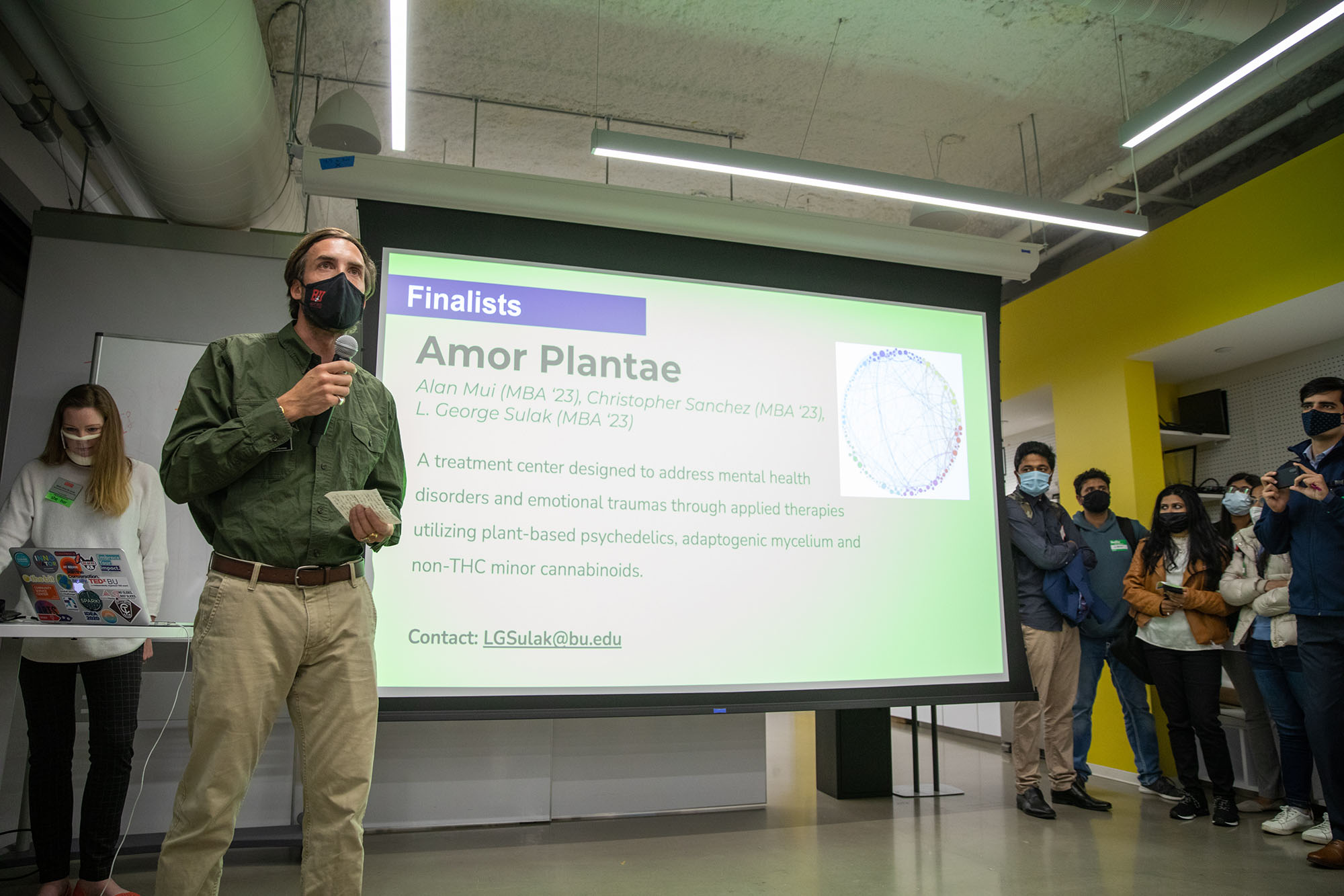 Photo of mask-wearing L. George Sulak, a first year MBA candidate for Social Impact (Questrom’23) talks about his winning team’s venture Amor Plantae, a treatment center used to treat mental illness through the use of plant-based psychedelics during Innovate@BU’s second annual Community Impact Challenge. He wears a green collared shirt and khaki pants as he stands in front of a large projector screen showing a slide for his project.