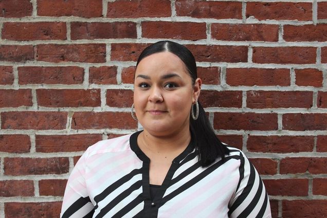 Photo of Angelica Rodriguez, with a white blouse with black stripes on it. She smiles slightly and stands in front of a brick wall.