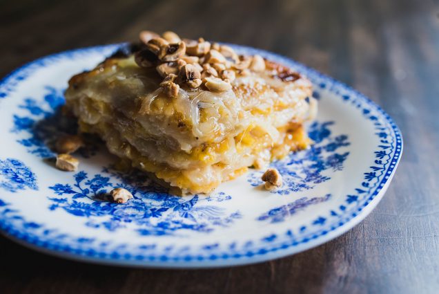 Photo of a slice of Butternut Squash and Potato Gratin topped with roasted hazelnuts on a white plate with ornate blue design. Cheese oozes from the gratin.