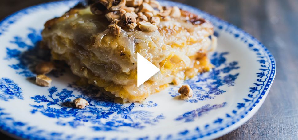 Photo of a slice of Butternut Squash and Potato Gratin topped with roasted hazelnuts on a white plate with ornate blue design. Cheese oozes from the gratin. White video play button is overlaid.