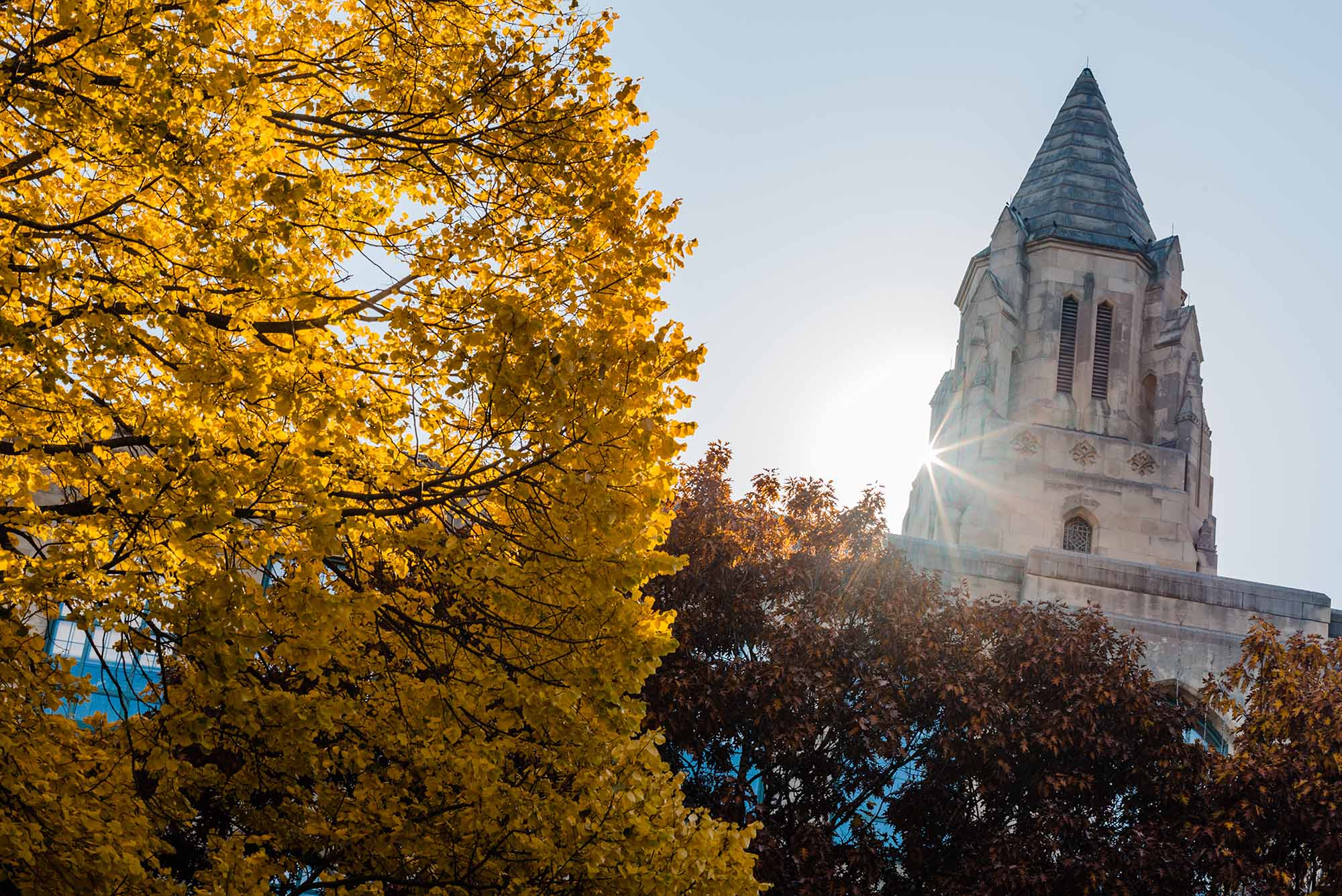 Photo of the top of an old church building. It is surrounded by Red and Yellow fall leaves as a sun glare peeks from around the top.