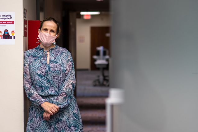 Dr Judy Platt leans on a classroom wall in while wearing a mask and a blue dress