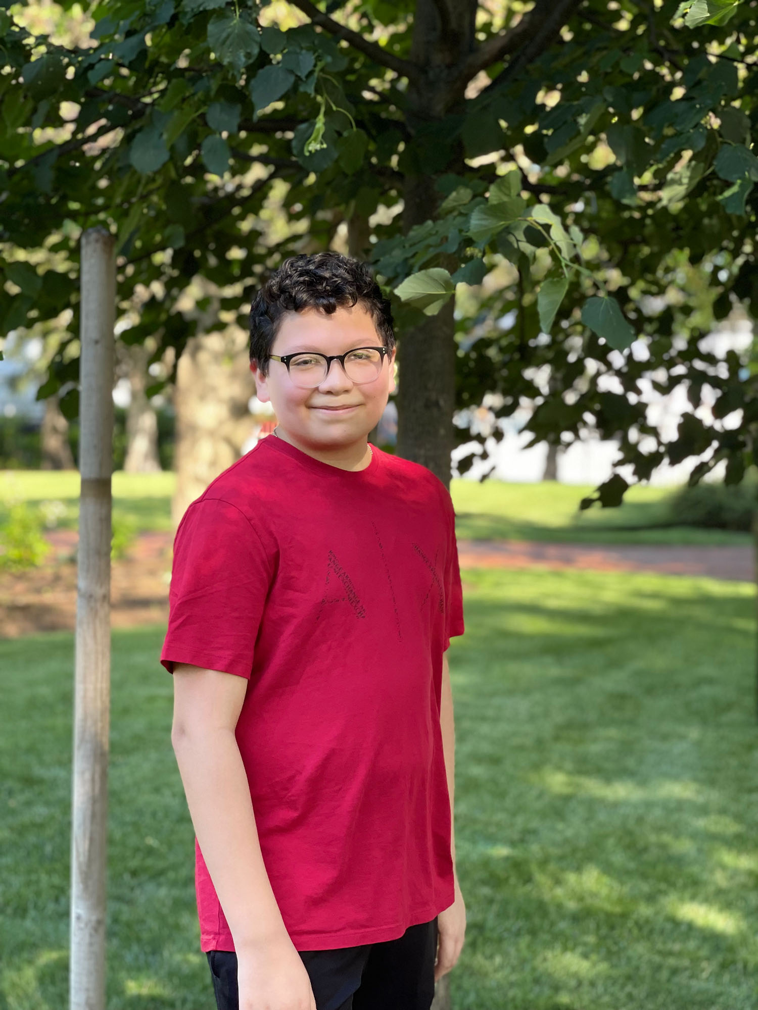 Photo of Steven Lue, a young boy with dark hair, black glasses, a red t-shirt. He stands in front of a small tree on a sunny day.