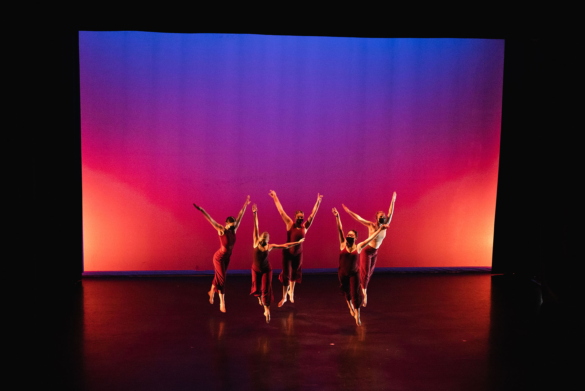 photo of 5 dancers on stage int he Fall 2021 Origins performance. They dance with toes pointed and arms in air, as they all wear black leotards and hair styled in sleek buns. They are illuminated by a red, purple, orange, and blue light that mimics the look of a sunrise on the backdrop.