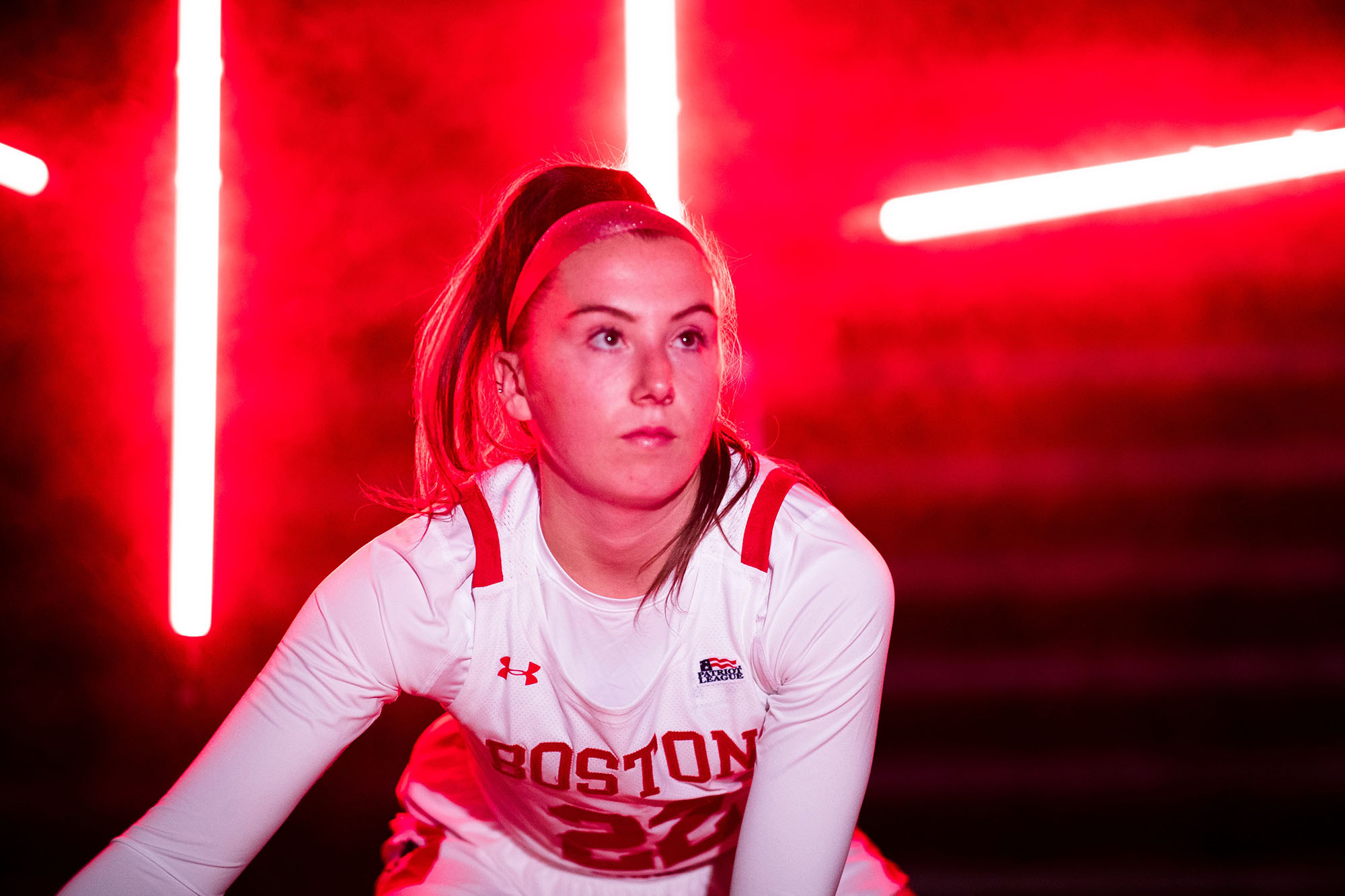 Action shot of Maggie Pina in her basketball uniform looking off to the distance as she dribbles a basketball. She's in a dark room lit by red bar lights giving off a misty red glow.