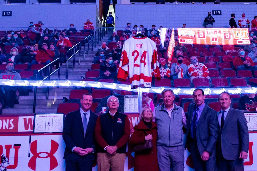 Photo of, from left to right, Albie O’Connell (CAS’99), current men’s hockey head coach; Jack Parker (Questrom’68, Hon.’97), former head coach; and Travis’s parents, Brenda and Lee at a memorial in the Agganis Arena for Travis Roy. The group stands at the edge of the ice, with a Travis Roy jersey hanging above them. Behind them, people sit in the stands.