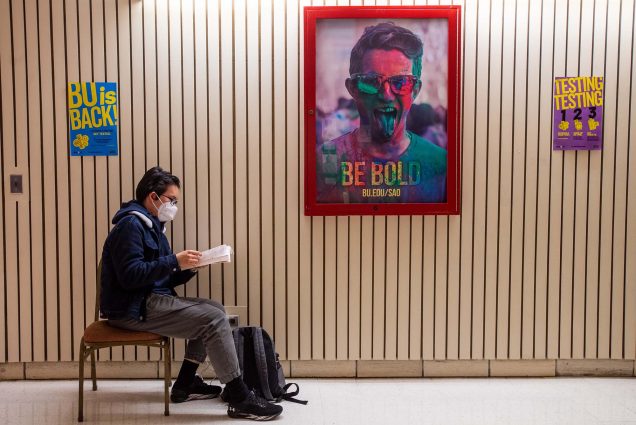 Photo of Jiangbei “Will” Li (CAS’22), wearing a white face mask and glasses, studying for his intro to Japanese course in the second floor hall of the GSU. He sits on a chair and charges his phone. A poster with the words "BE BOLD" hangs nearby and shows a student covered in colored paint.
