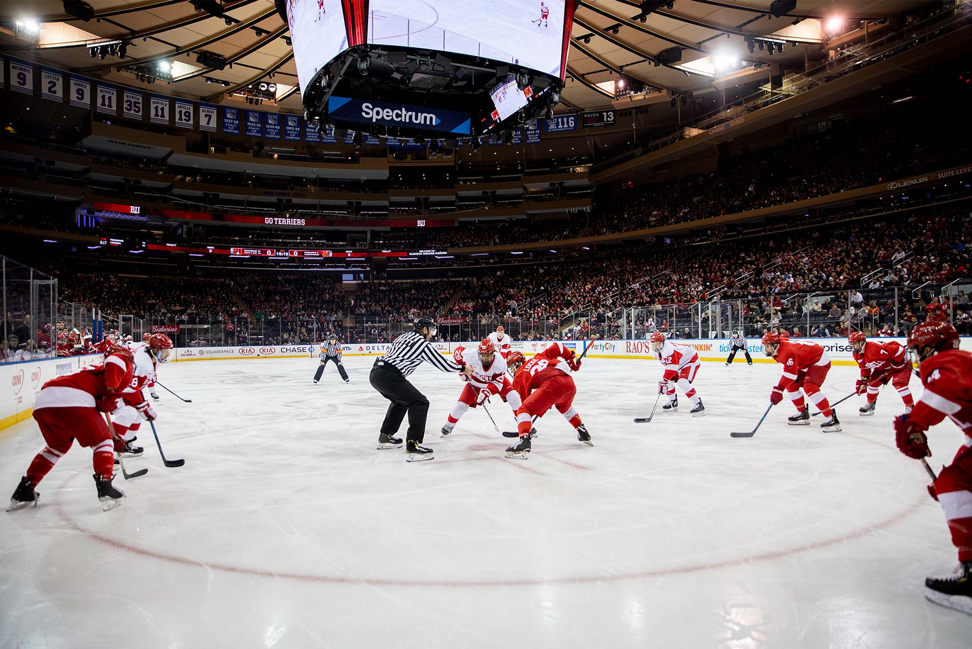 photo of the Bu men's hockey team mid-puck drop facing Cornell. Stands are crowded and both teams stand poised on the ice as a ref stands at ready.