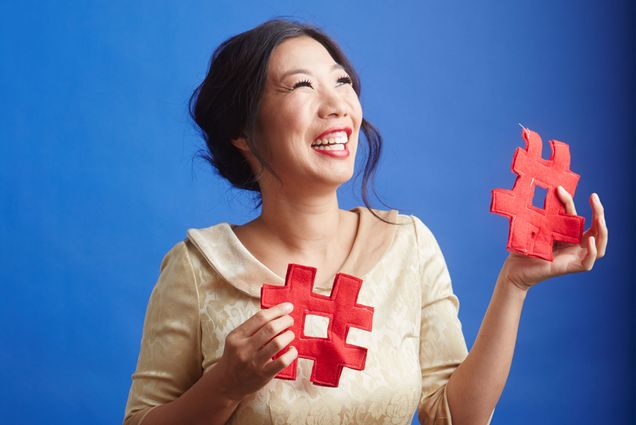 Kristina Wong stands looking upward with a smile on her face. She is holding 2 red pound symbols that appear to be made of felt.