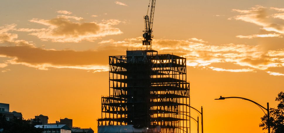 photo of BU’s new 19-story Center for Computing & Data Sciences during construction. A crane is atop the structure. It serves as a looming figure during a scenic orange sunset. Cars are seen in the horizon.