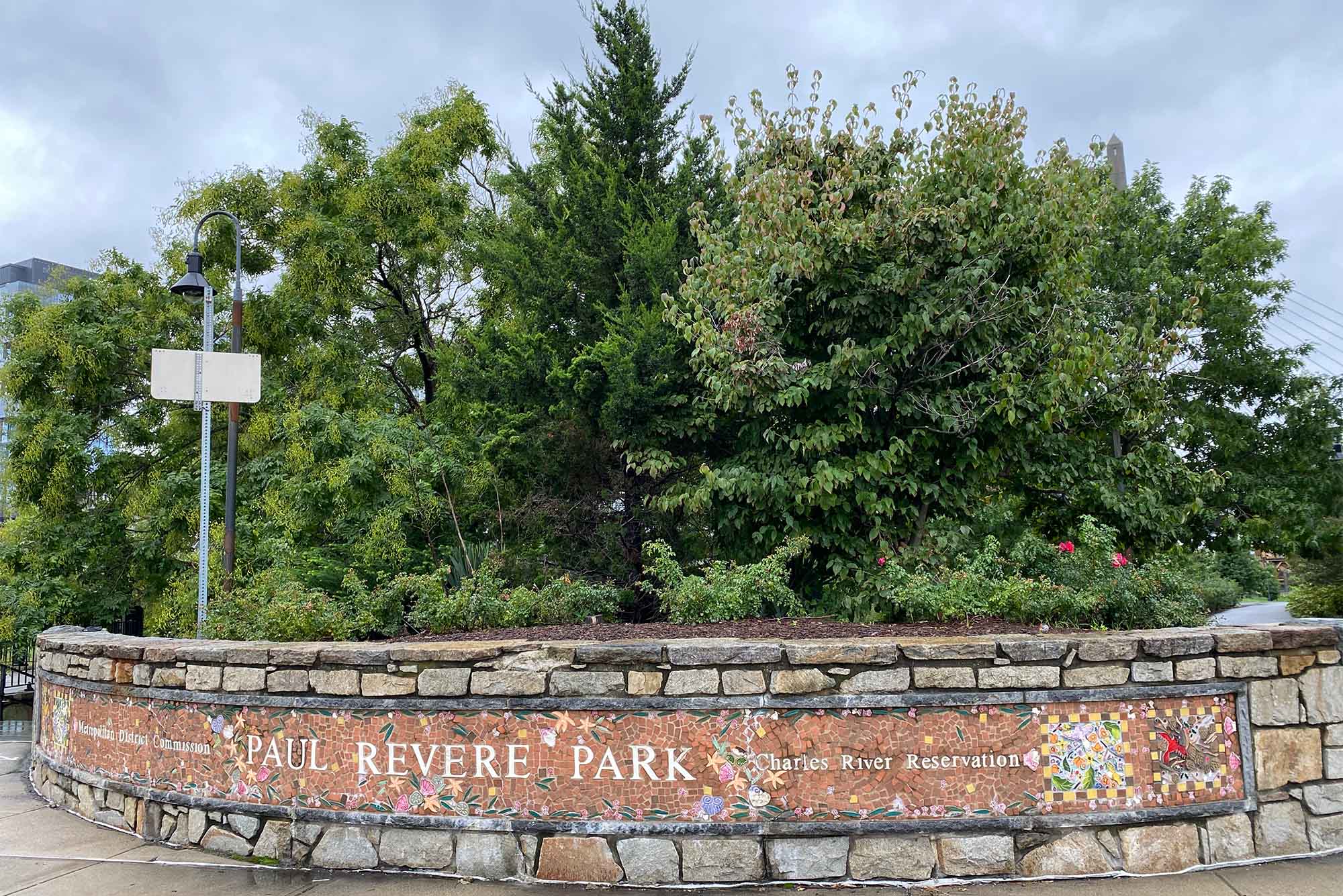 A sign reads "Paul Revere Park" in stone in front of a backdrop of evergreen trees