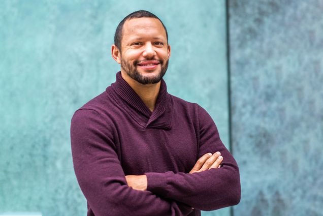 Doug Densmore poses with arms crossed for a photo in front of a light blue wall. He wears a burgundy sweater and smiles at the camera.