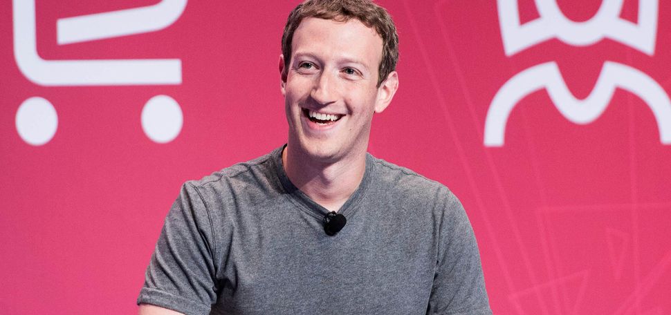 photo of CEO Mark Zuckerberg announcing that Facebook is changing its corporate name to Meta. Smiling and wearing a grey shirt, he looks to the left and laughs. He sits in front of a rose-colored background with white icons of a shopping cart and female person behind him.
