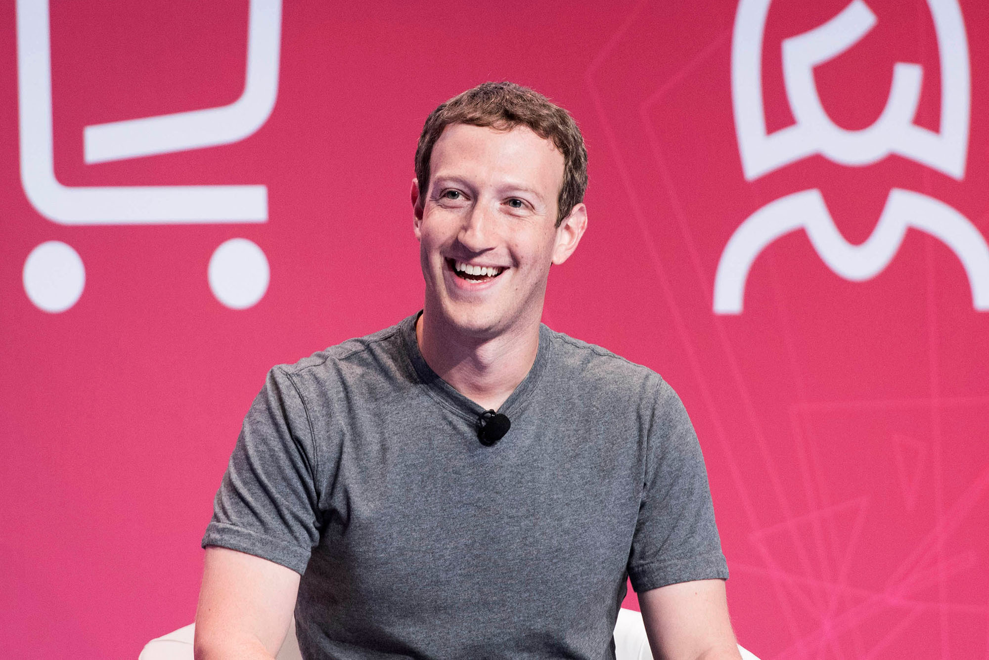 photo of CEO Mark Zuckerberg announcing that Facebook is changing its corporate name to Meta. Smiling and wearing a grey shirt, he looks to the left and laughs. He sits in front of a rose-colored background with white icons of a shopping cart and female person behind him.