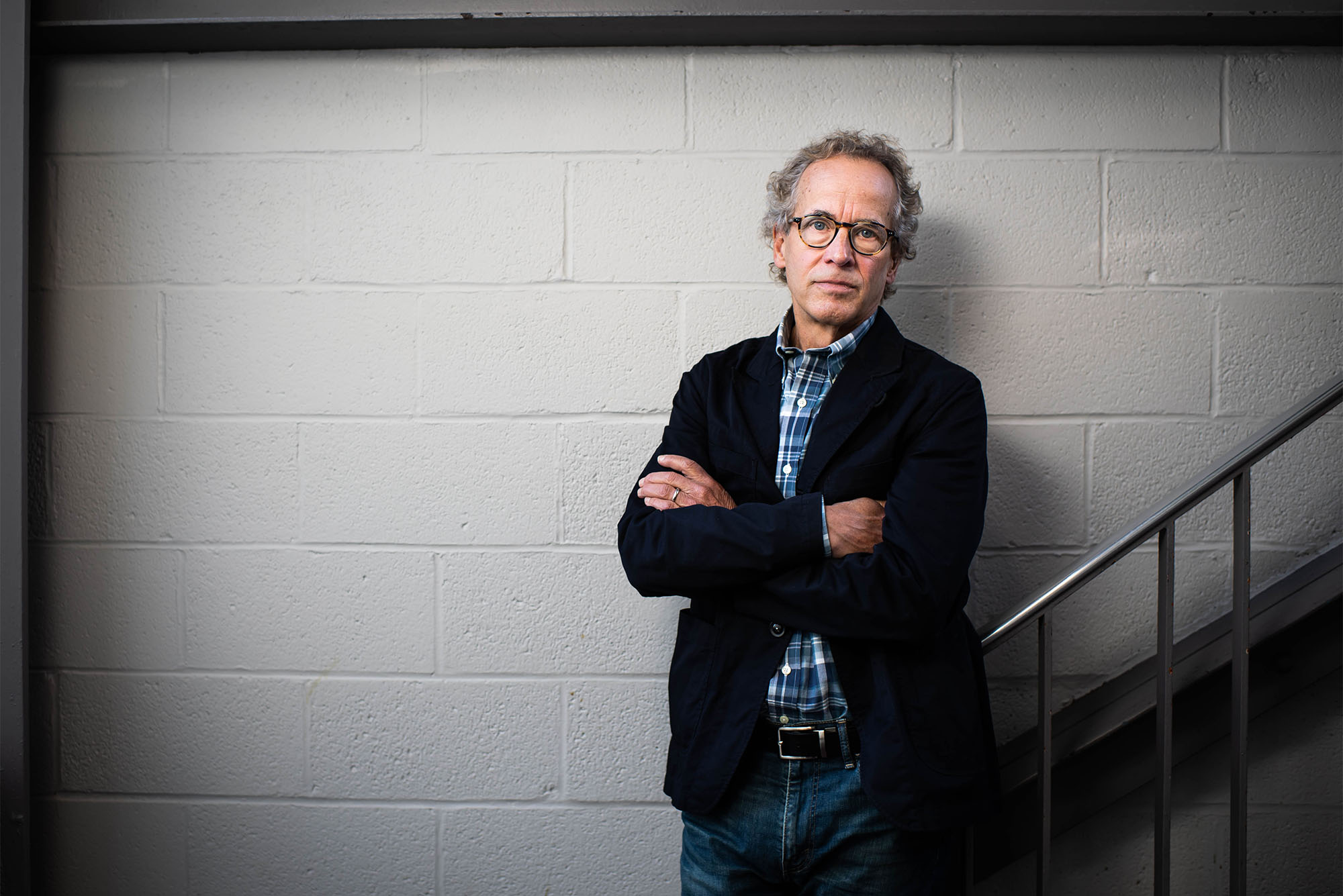 Dick Lehr stands in front of a white cinderblock wall wearing a suitcoat and glasses with his arms crossed