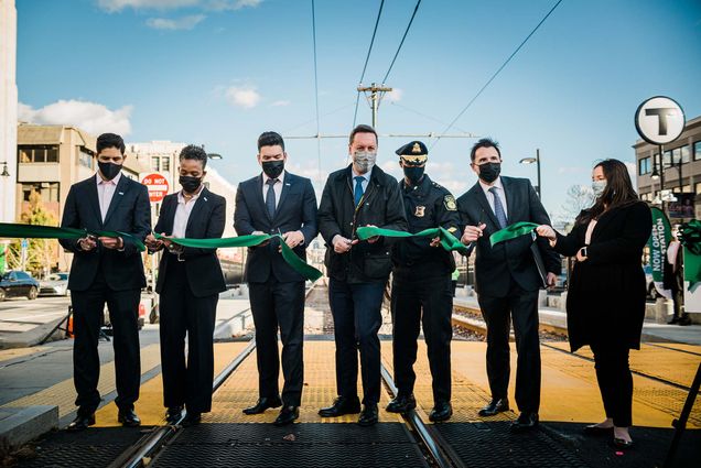 Photo of a group of MBTA officials dressed in dark suits and/or formal attire and wearing dark masks as they cut a green ribbon. They stand on the green line track and blue sky is seen behind them. From left to right: Andres Achury – GLT Senior Director Desiree Patrice – Deputy Chief, Angel Peña MBTA Chief Capital Transformation Steve Poftak – MBTA General Manager Kenneth Green – Chief Transit Police Derek Howe – BU Senior Vice President of Operations Shauna Connelly—GLT Senior Project Coordinator. Photo by Janice Checchio