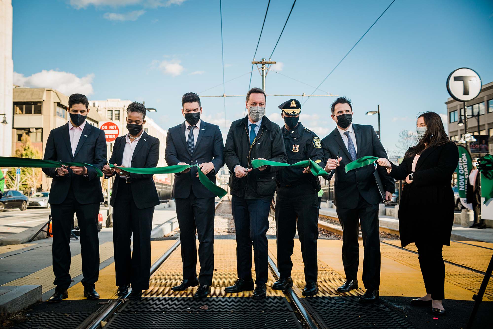 Photo of a group of MBTA officials dressed in dark suits and/or formal attire and wearing dark masks as they cut a green ribbon. They stand on the green line track and blue sky is seen behind them. From left to right: Andres Achury – GLT Senior Director Desiree Patrice – Deputy Chief, Angel Peña MBTA Chief Capital Transformation Steve Poftak – MBTA General Manager Kenneth Green – Chief Transit Police Derek Howe – BU Senior Vice President of Operations Shauna Connelly—GLT Senior Project Coordinator. Photo by Janice Checchio