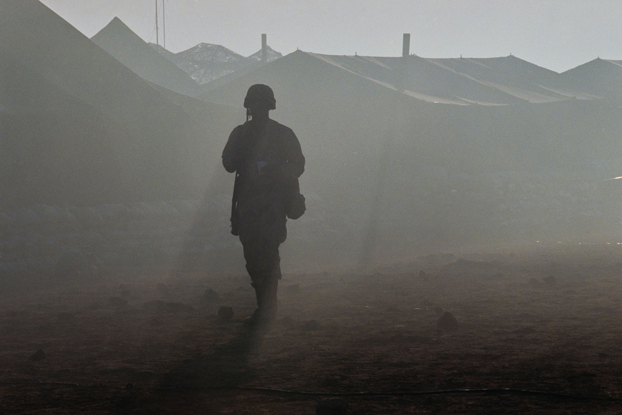 Photo of a silhouette of a soldier in military fatigues, with the tops of the tents in a military camp visible through the desert dust in the background, in the Saudi–Iraqi neutral zone on the border between Saudi Arabia and the Iraq, 1991.