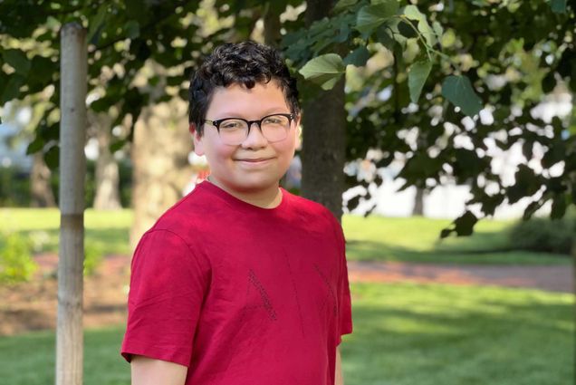 Photo of Steven Lue, a young boy with dark hair, black glasses, a red t-shirt. He stands in front of a small tree on a sunny day.