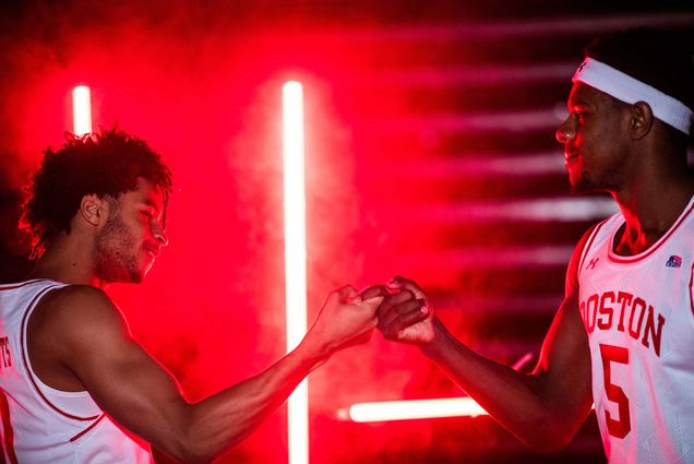 Photo of teammates Ethan Brittain-Watts (left) (CAS’23) and Walter Whyte (right) , two young Black men, clasping fingers as they smile. They both wear white home jerseys and red colored smoke is seen behind them.