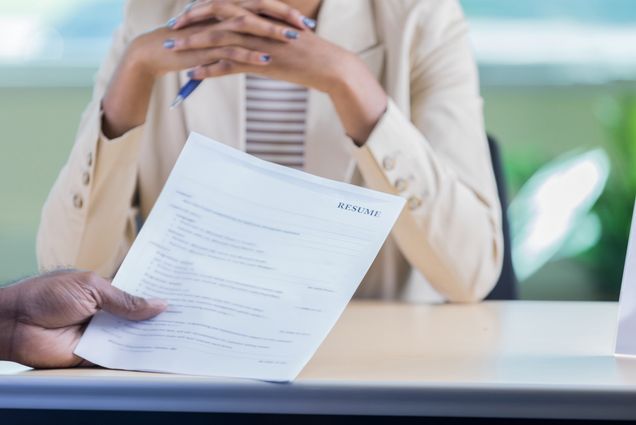 Unrecognizable businessperson conducts a job interview with an unrecognizable female candidate. One hand is holding the woman's resume. The woman is sitting across the desk from them. Photo via iStock / SDI Productions