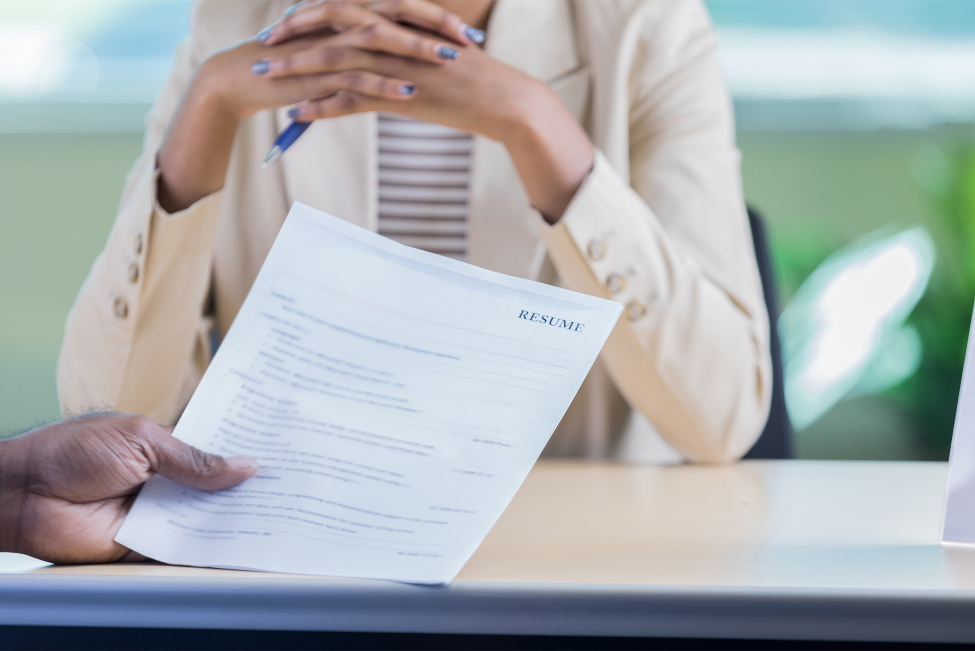 Unrecognizable businessperson conducts a job interview with an unrecognizable female candidate. One hand is holding the woman's resume. The woman is sitting across the desk from them. Photo via iStock / SDI Productions