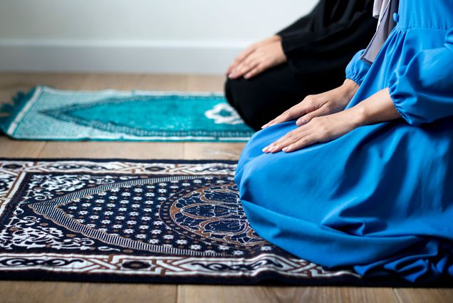 Photo of two Muslim women worshiping on prayer rugs. They kneel on the rugs that are decorated in ornate patterns.