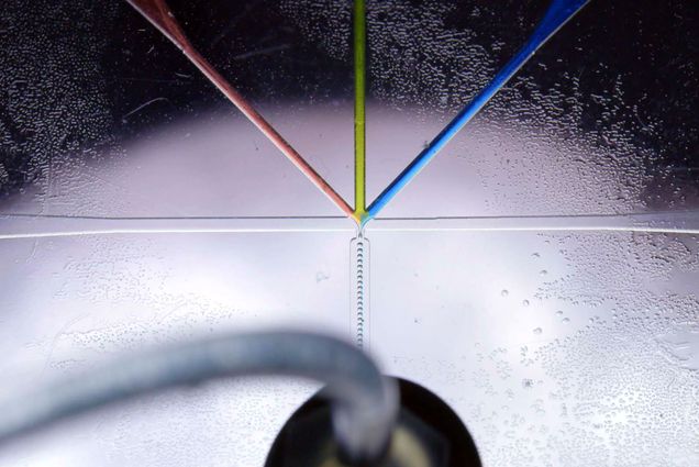 Image of a microfluidic device, a tiny circuit boards for analyzing liquids, running. a red, yellow, and blue liquid come together from 3 different to meet in the center and combine to create a new chemical that streams through a new tube in tiny droplets