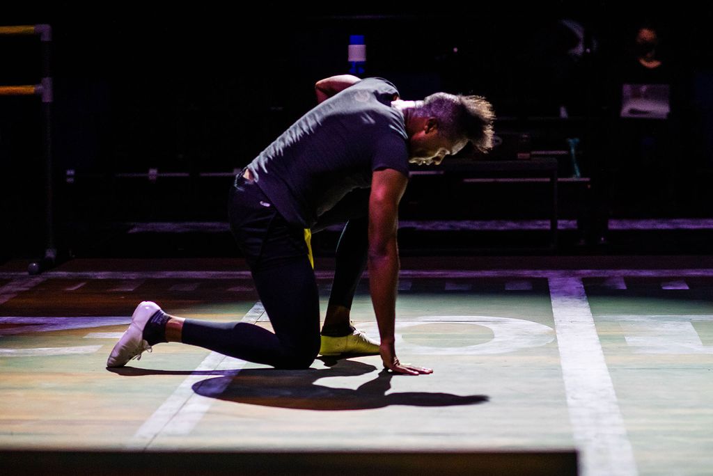 Photo of Donovan Black (CFA 22), a young, tall, Black man, wearing a gray t-shirt and shoes similar to ballet slippers, while rehearsing for Colossal. He kneels center stage and puts his hand on the ground.