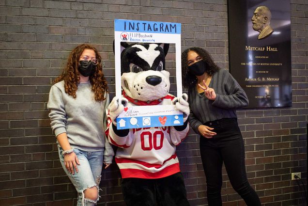 Photo of Genesis Belasco (SAR 24) (left) and Sophia Cornish taking a photo with Rhett during a First-Gen College Celebration on Monday, November 8, 2021. Rhett holds a frame around their face that makes them look like an instagram post. The girls smile and wear facemasks.