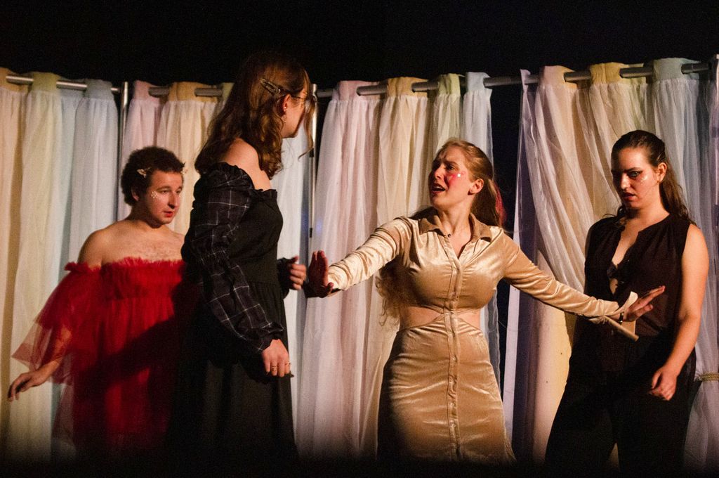 Photo of a rehearsal featuring: Ian Aquino (COM’23) as Helena, Ella Moran (CAS’25) as Lysander, Carolyn “Carrie” Welter (CAS’22) as Hermia, and Emma Permé as Demetrius. Welter wears a shiney gold dress and stands between two characters who look like they may be about to fight.