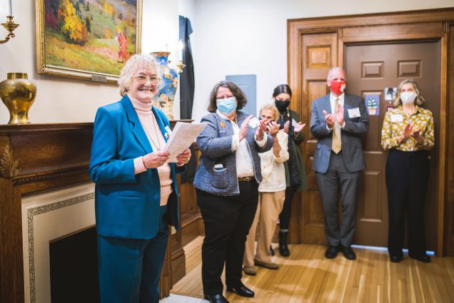 Photo of Diane Gallagher, left, in a bright blue blazer, holding a piece of paper, as she speaks during the Fisk House dedication on Friday, November 5, 2021. Other masked members of the BU community are seen behind her uploading; they stand in a room with wooden floors and wood panelling.