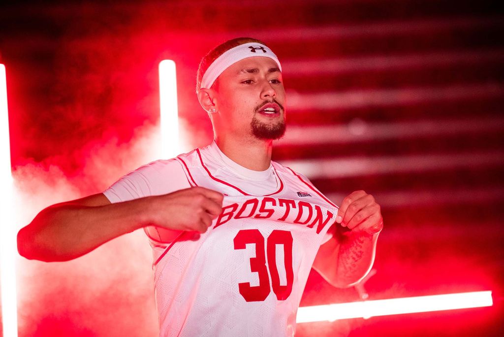 Photo of Javante McCoy (Questrom’21, SHA’22) pulling on the top of his white jersey with the number 30 and "Boston" printed on it in red. He wears a white under armor headband and red smoke is seen behind him.