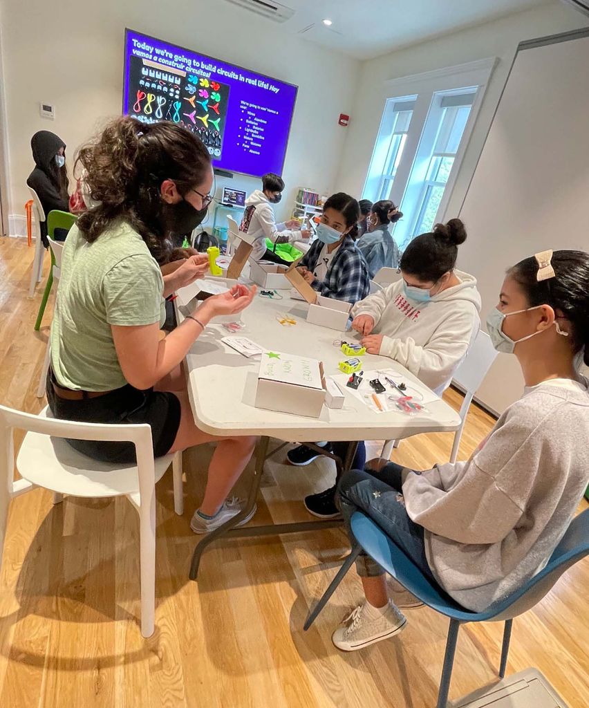 Photo of Michalina (Mica) Jadick (green shirt at left), one of the BU STEM undergraduates who worked part of the team with Paul Trunfio on co-designing the Network Science curriculum. In the photo she is supporting the youth in her group to create their own circuits to light the light bulb. Mica  sits at a low, white table with a group of young people as they complete an activity. Everyone wears face masks.